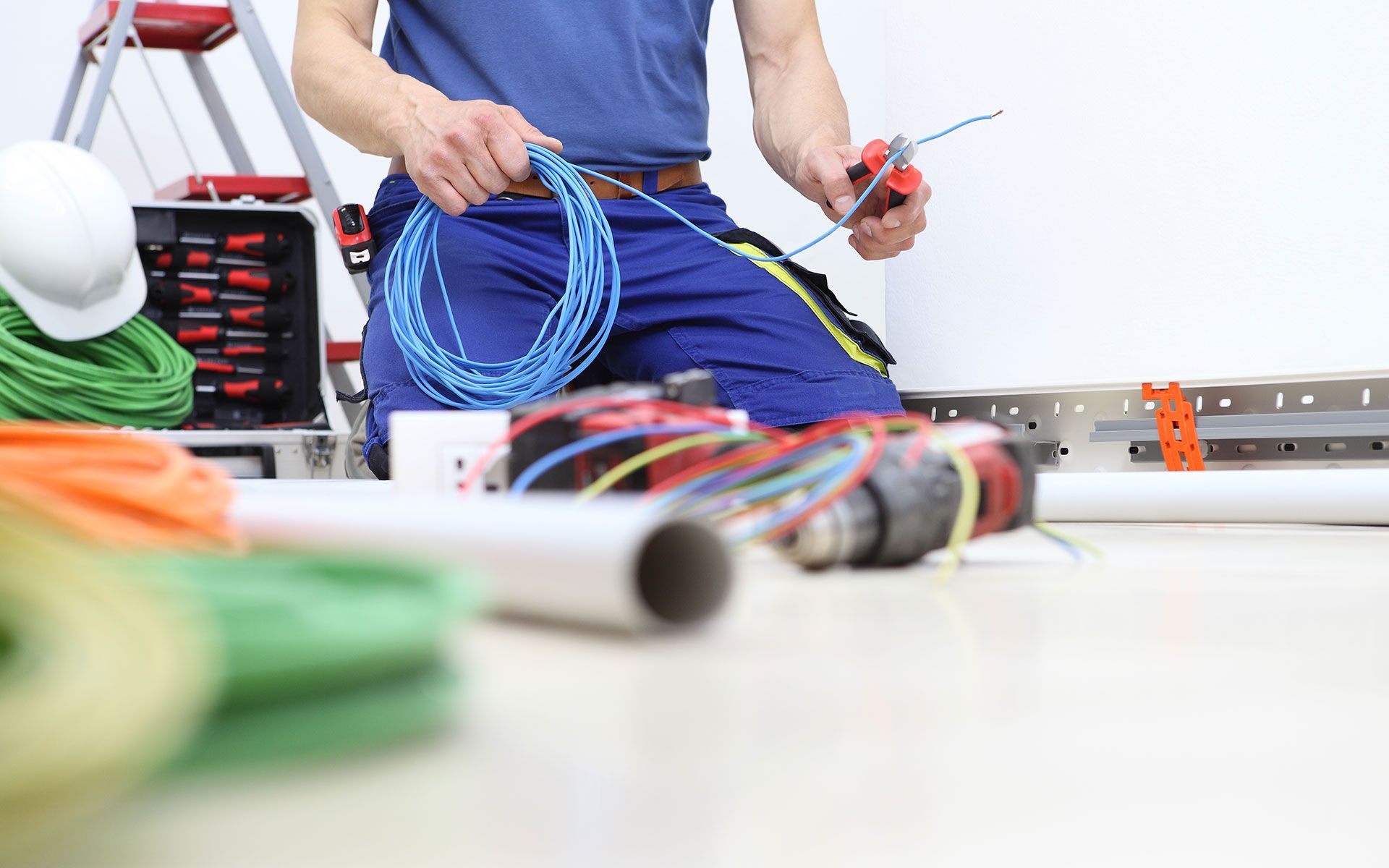 Electrician kneeling, cutting blue wire with pliers, surrounded by wires and tools.