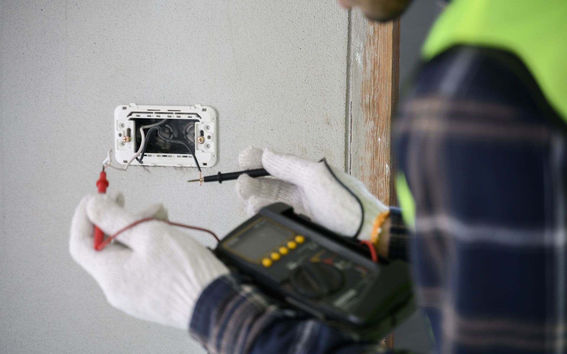 Electrician testing wires in a wall outlet with a multimeter, wearing gloves and a safety vest.