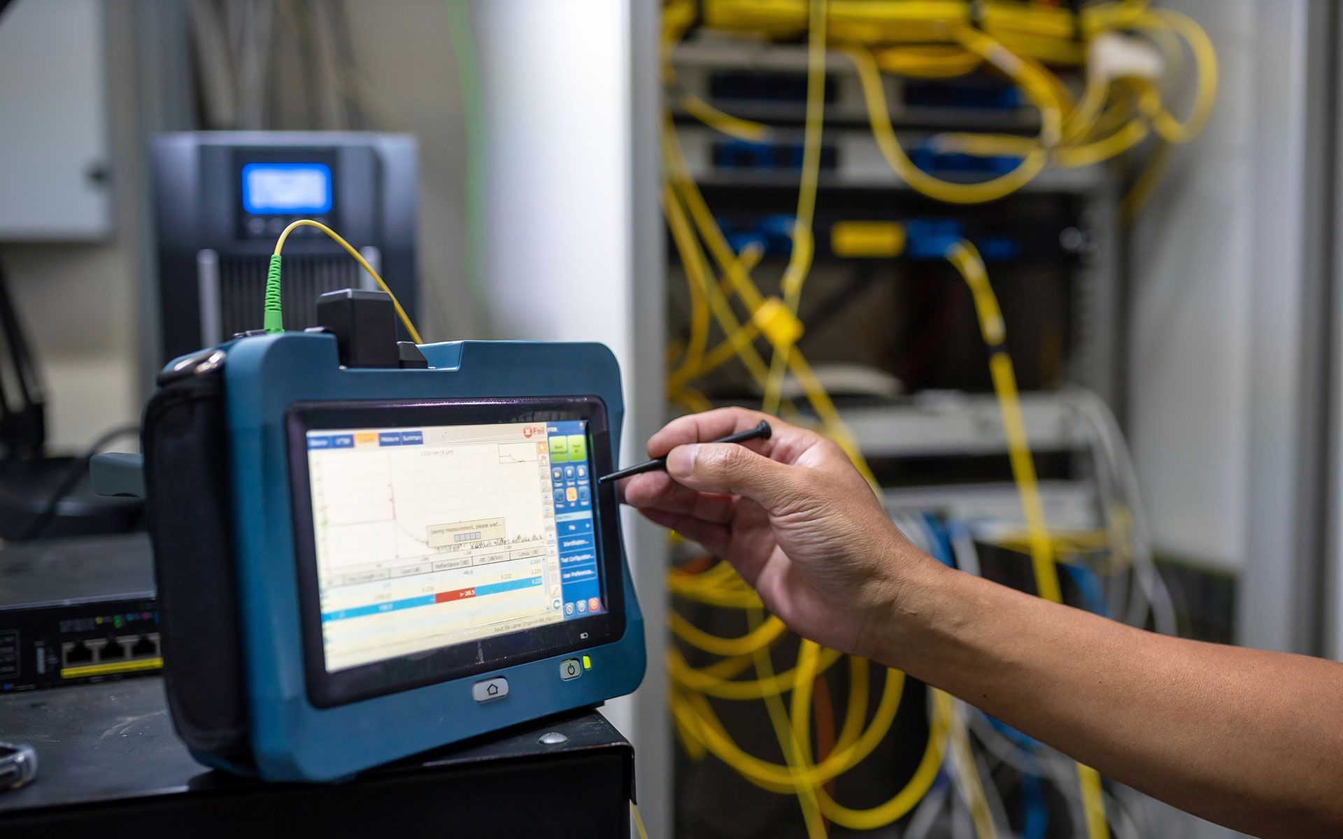 Person using a device to test fiber optic cables in a server room. The device displays a graph on its screen.