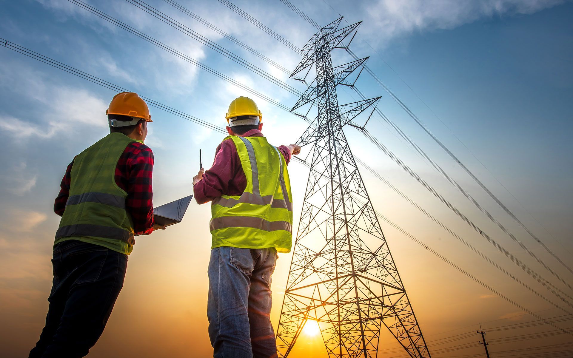 Two workers in safety vests and hard hats examining power lines and a transmission tower at sunset.