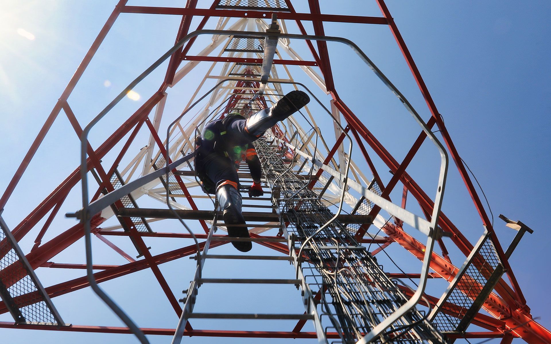 Person climbing a tall red and white communications tower on a sunny day.