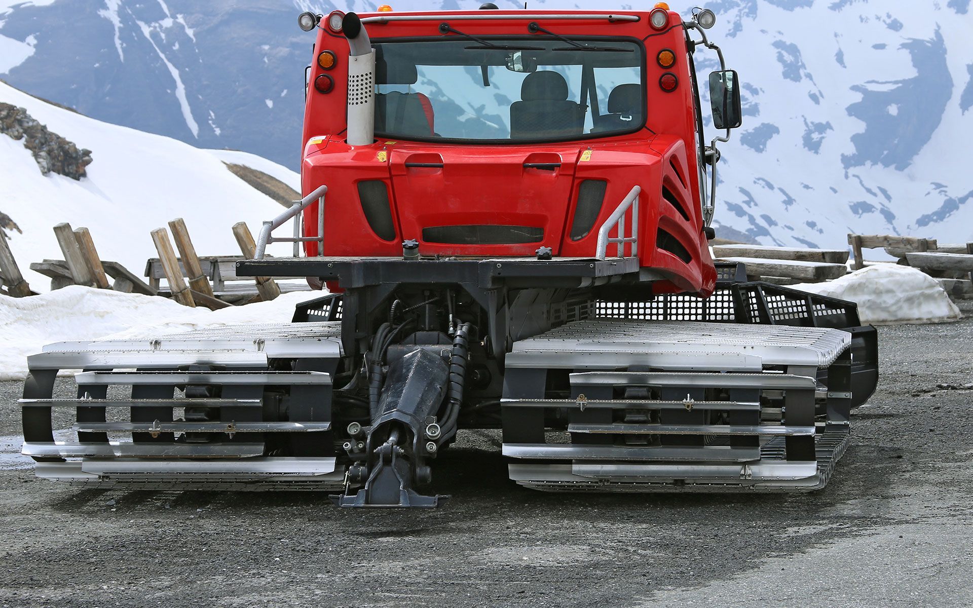 Red snow groomer on gravel road, with tracks, near snow-covered mountain.