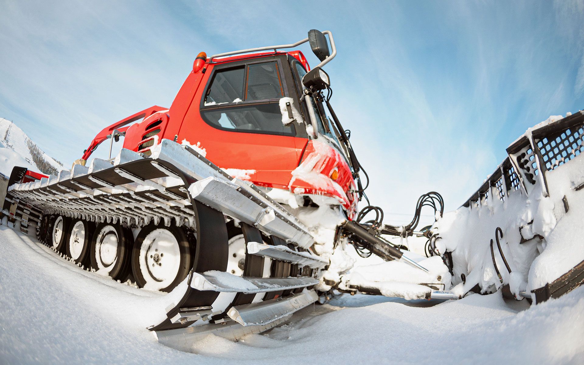 Red snow groomer on snow, tracks visible, under a blue sky.