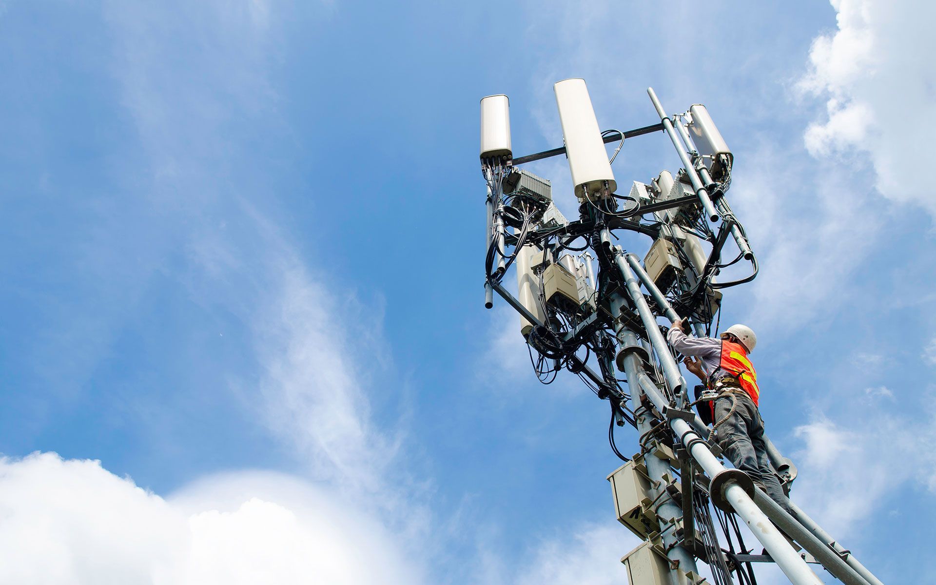 Worker on a telecommunications tower, adjusting equipment against a blue sky with clouds.