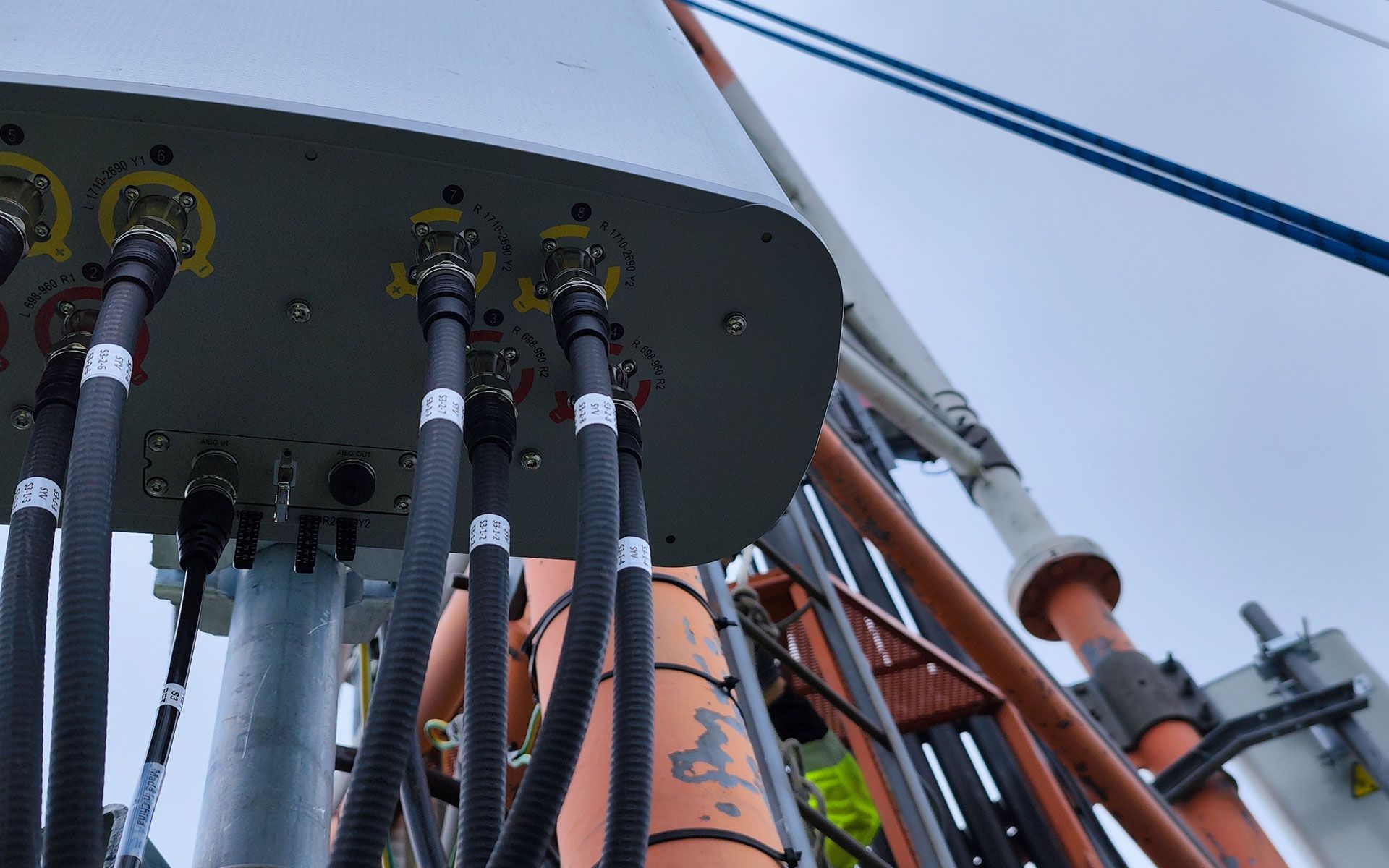 Close-up of gray device with multiple black cables and connectors, mounted on a tower against a cloudy sky.