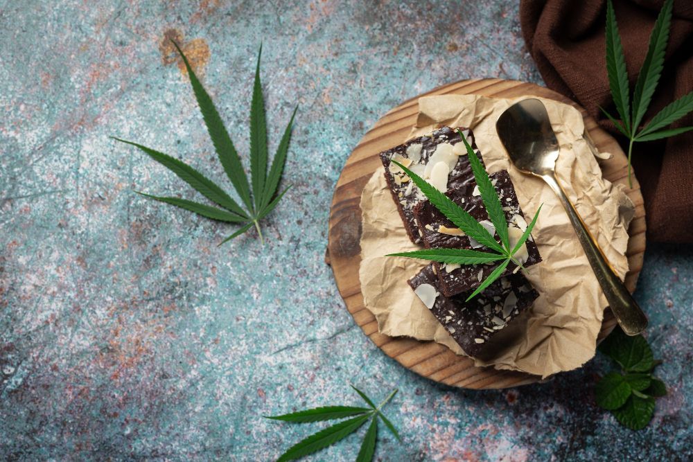 Marijuana edibles on a wooden plate with a spoon. Green cannabis leaves are scattered around the plate on a textured teal background.