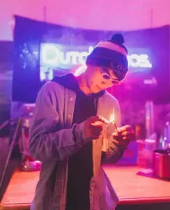 Person in a blue beanie and sunglasses lights a match, neon sign 