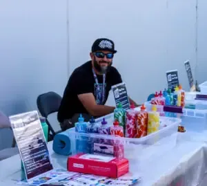 Man smiles at a table with colorful art supplies, possibly selling art. He wears sunglasses and a cap.