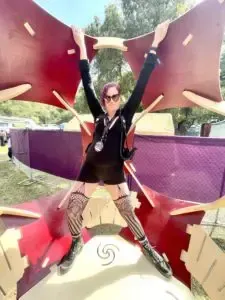 Woman in black dress and patterned stockings poses in front of a geometric art piece with arms raised.