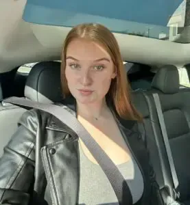 A young woman with auburn hair wearing a black leather jacket and seatbelt inside a car. She's looking at the camera.