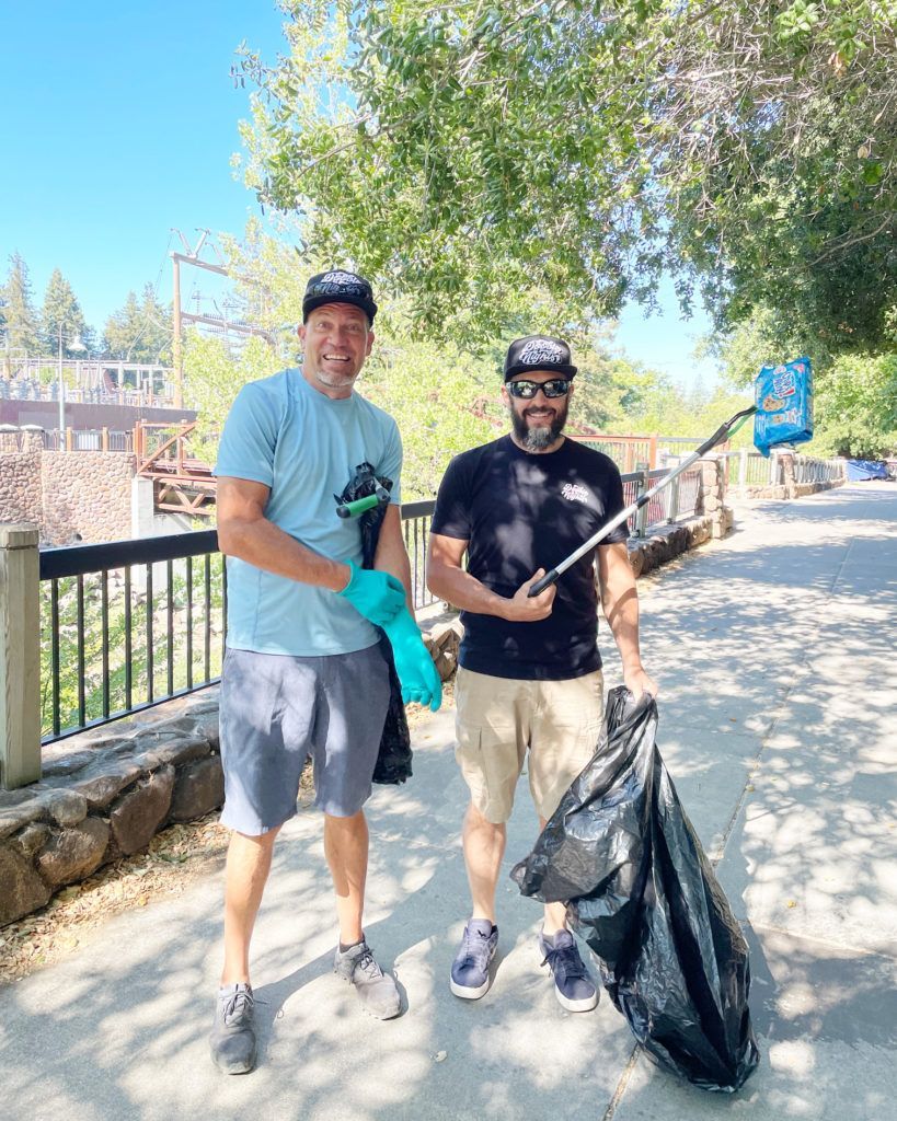 Two men smile, holding trash bags and grabbers, on a sunny day near a bridge and trees. They are collecting litter.