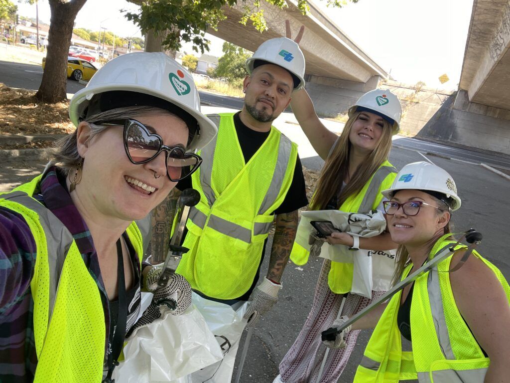 Four people in hard hats and safety vests take a selfie outdoors under a bridge. They are smiling and posing.