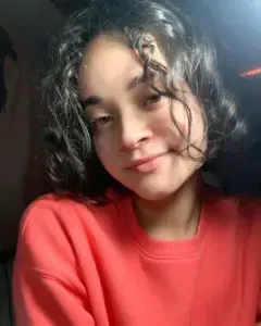 Young person with curly dark hair, wearing an orange sweater, smiling slightly.