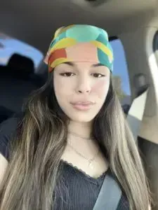 Woman with long brown hair wearing a colorful headscarf, black top, and necklace, in a car.