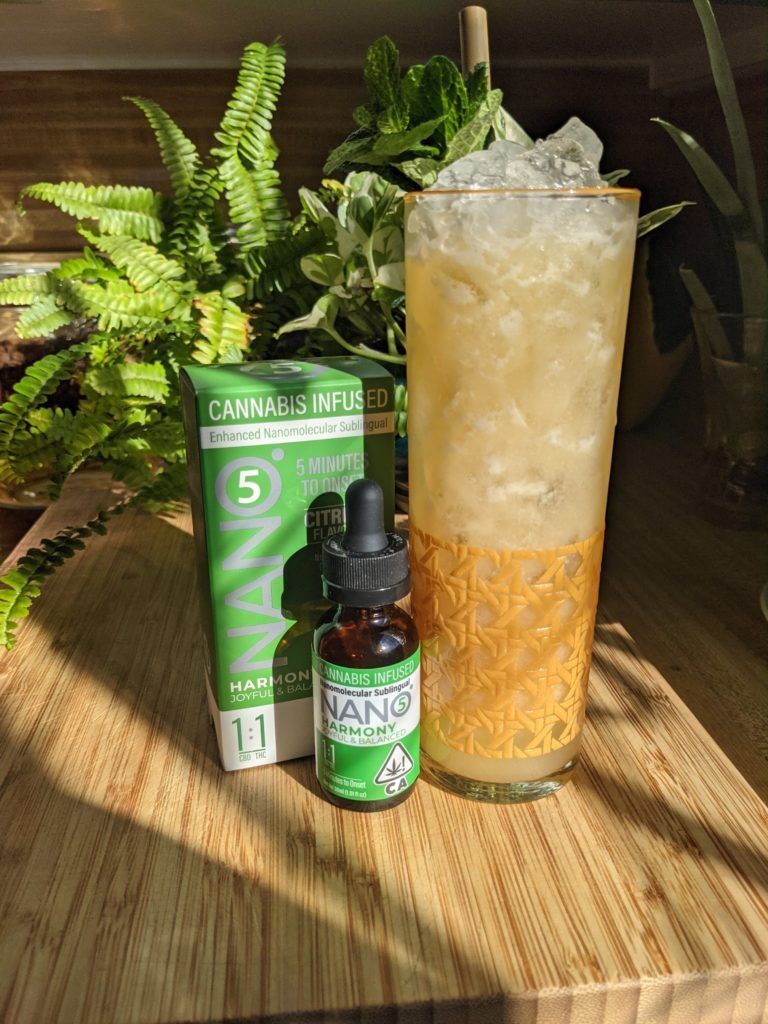A cannabis-infused beverage with dropper bottle and box, on a wooden surface with plants.
