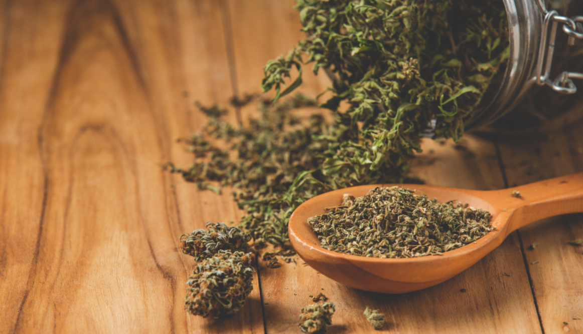 A jar of cannabis spilling out onto a wooden table, with nugs and a spoonful of shake next to it.