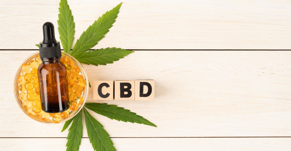 A bowl of gelcaps with a tincture bottle on top, on a light table with the wooden letter tiles CBD next to it.