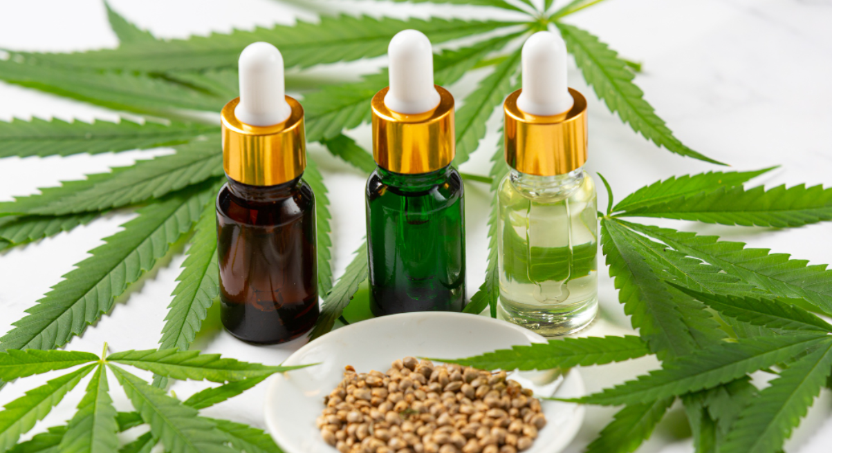 Three tincture bottles on a white surface with cannabis leaves and a bowl of cannabis seeds in front of them.