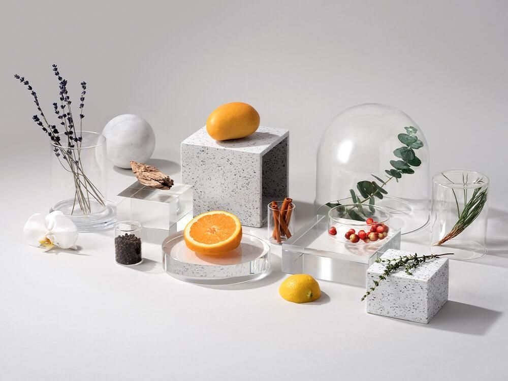Assortment of ingredients like fruit, herbs, and spices artfully arranged on pedestals, with vases on a white surface.