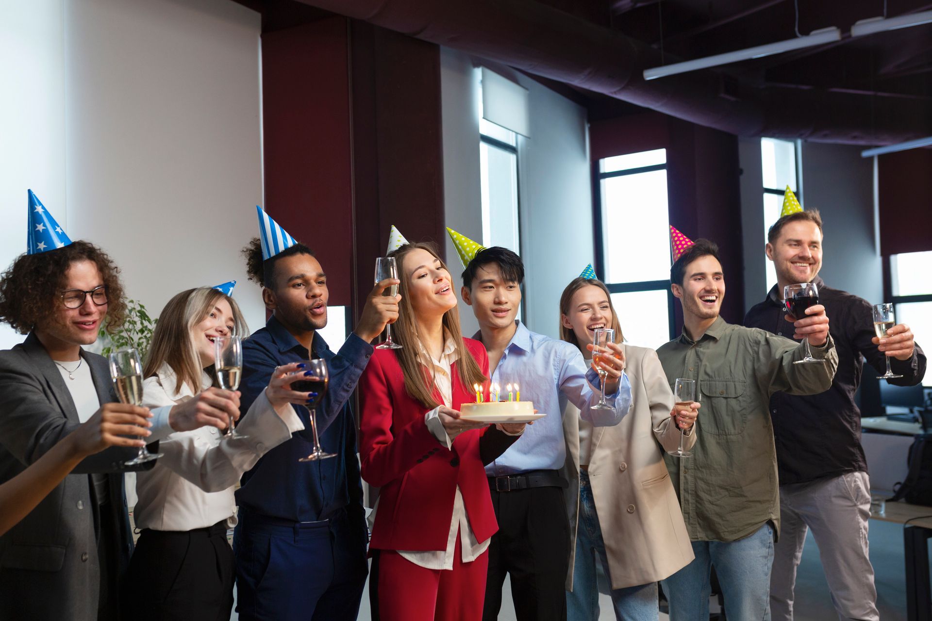 A group of colleagues in colorful birthday hats sing and toast at a birthday party.