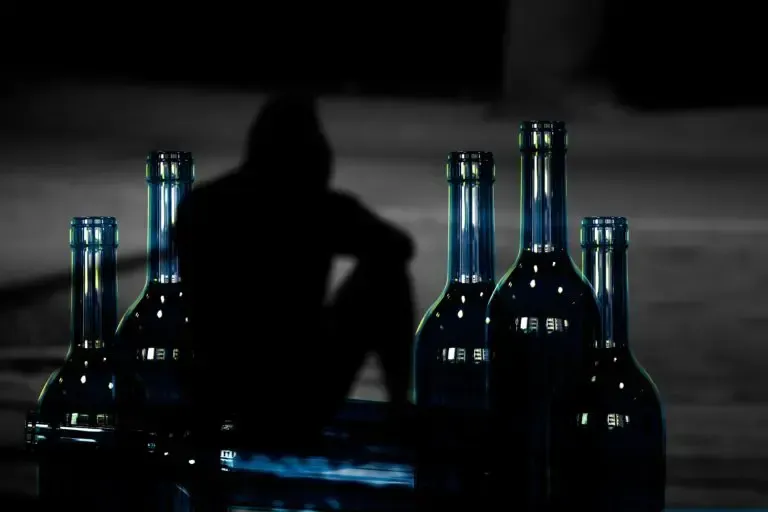 Silhouette of a person sitting near empty bottles, likely suggesting substance abuse. Dark setting.