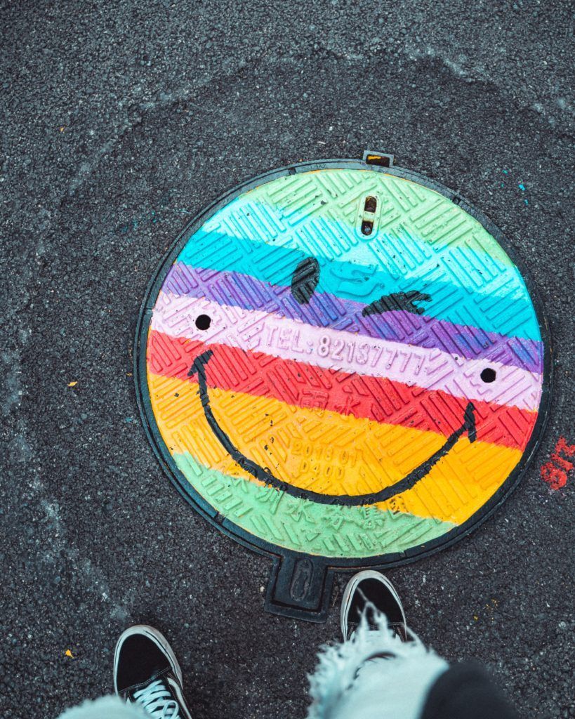 Manhole cover painted with a colorful, winking smiley face on a textured gray surface.
