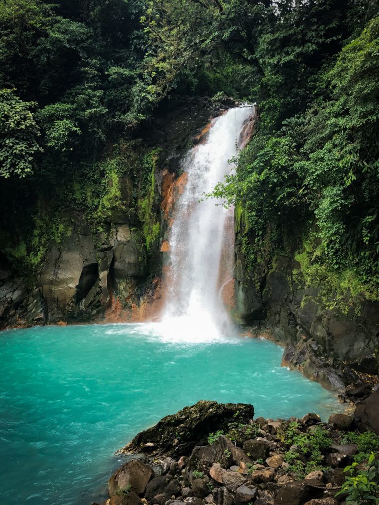 Waterfall cascading into a turquoise pool surrounded by lush green forest and rocky terrain.