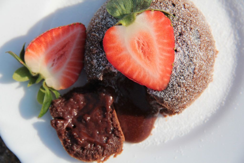 Chocolate lava cake with strawberries on a white plate. Molten chocolate oozes out; the cake is dusted with powdered sugar.