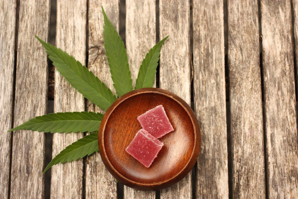 Two pink, square gummies in a wooden bowl, next to a cannabis leaf, on a rustic wooden surface.