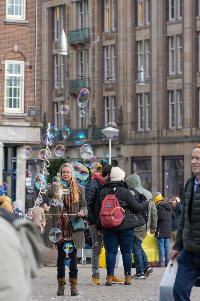 Woman blowing bubbles in a city square, surrounded by people. Tall buildings in the background.
