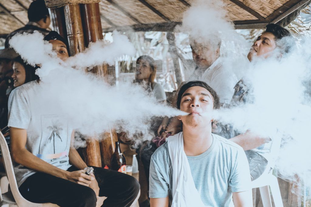 Group of people exhaling large clouds of vapor. The central figure is blowing a cloud directly at the viewer.