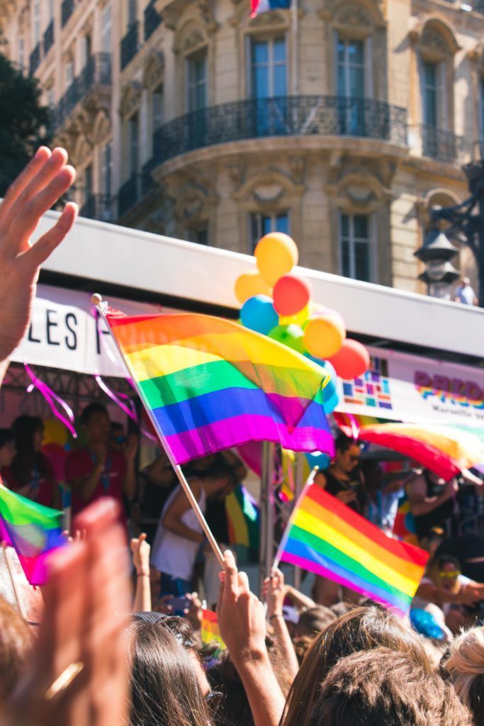 People at a Pride parade wave rainbow flags, celebrating in a city setting. Balloons float above a crowd.