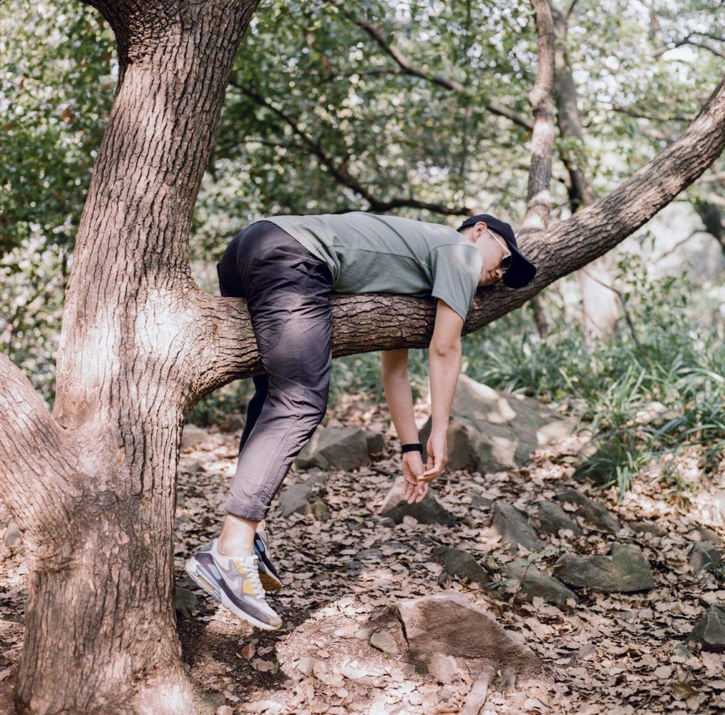 Man resting on a tree branch, wearing a hat, green shirt, and dark jeans, in a wooded area with light sunlight.