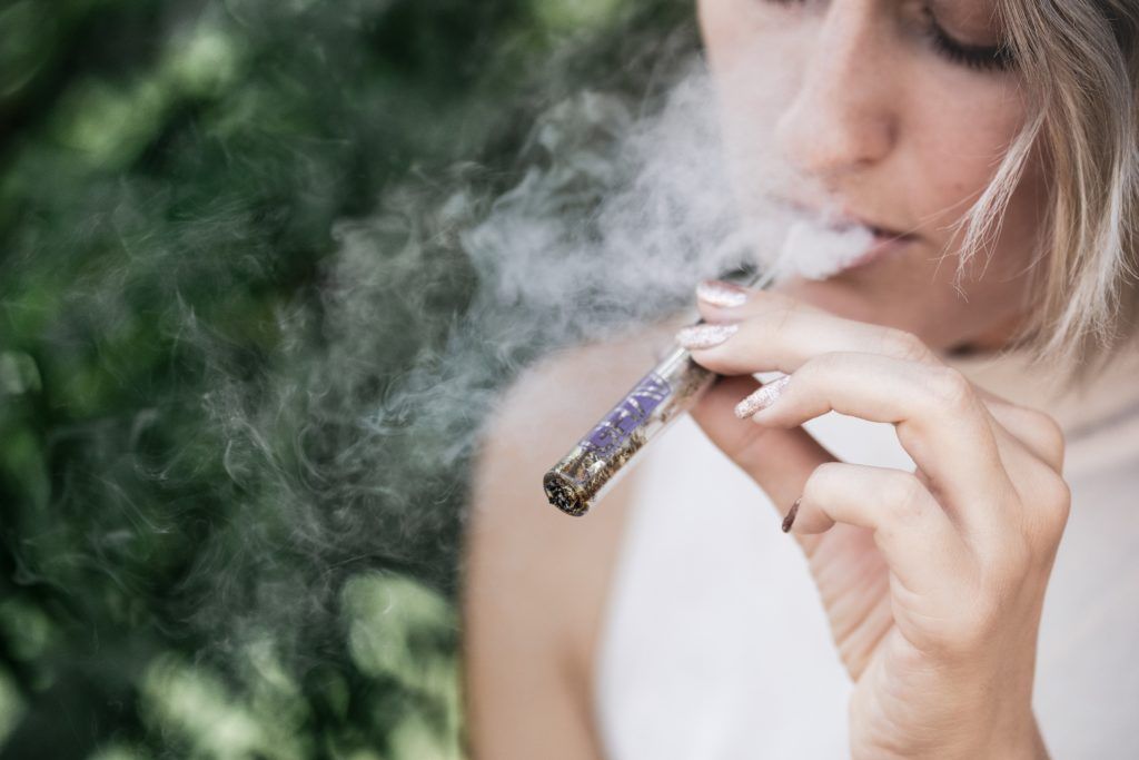 A woman exhales smoke from a lit joint, outdoors with blurred greenery in the background.