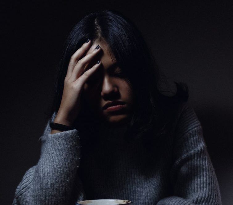 A person with dark hair and a gray sweater holds their head in their hand, appearing stressed in a dimly lit setting.