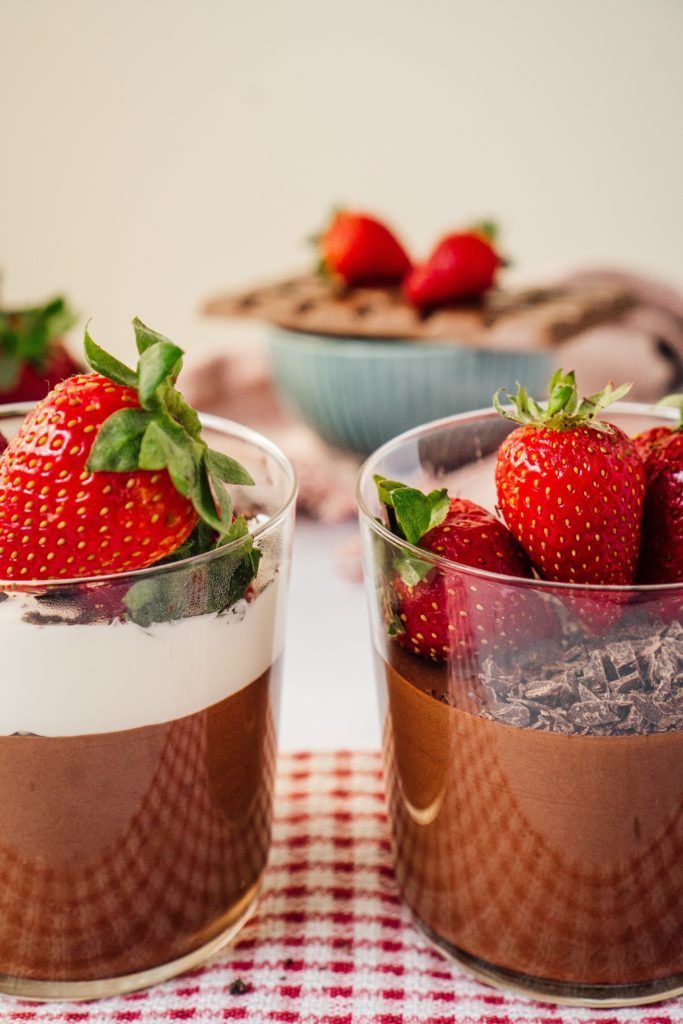 Chocolate dessert parfaits topped with strawberries in glass cups, set on a tablecloth with a bowl in the background.