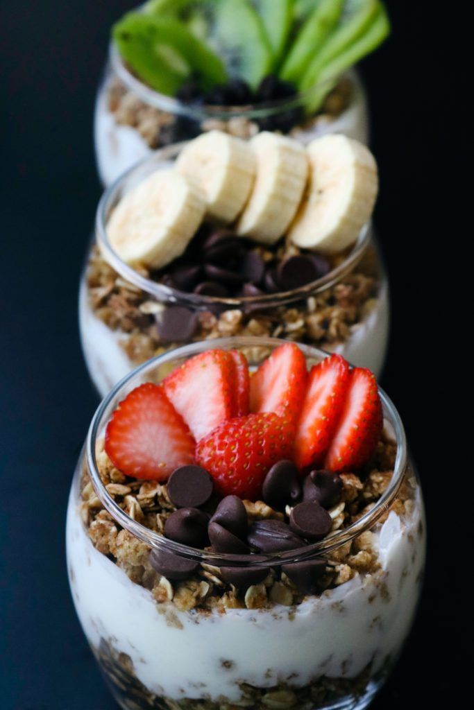 Three layered yogurt parfaits topped with fresh fruit and chocolate chips in glass cups on a black surface.
