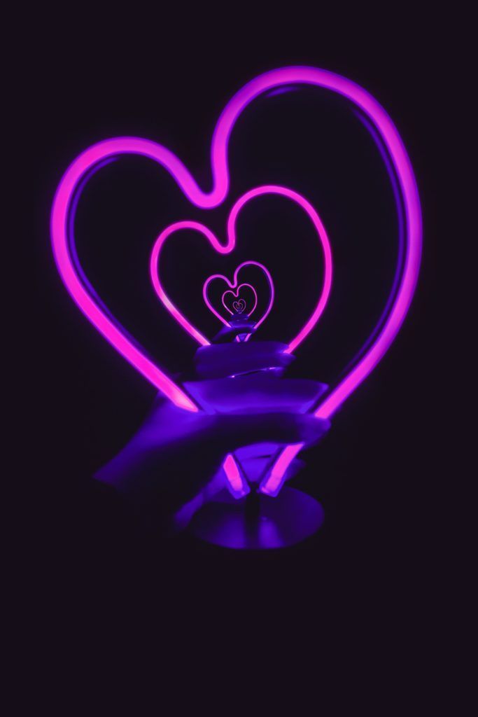 A hand holds a neon pink heart-shaped light against a black background, creating an infinite heart tunnel effect.
