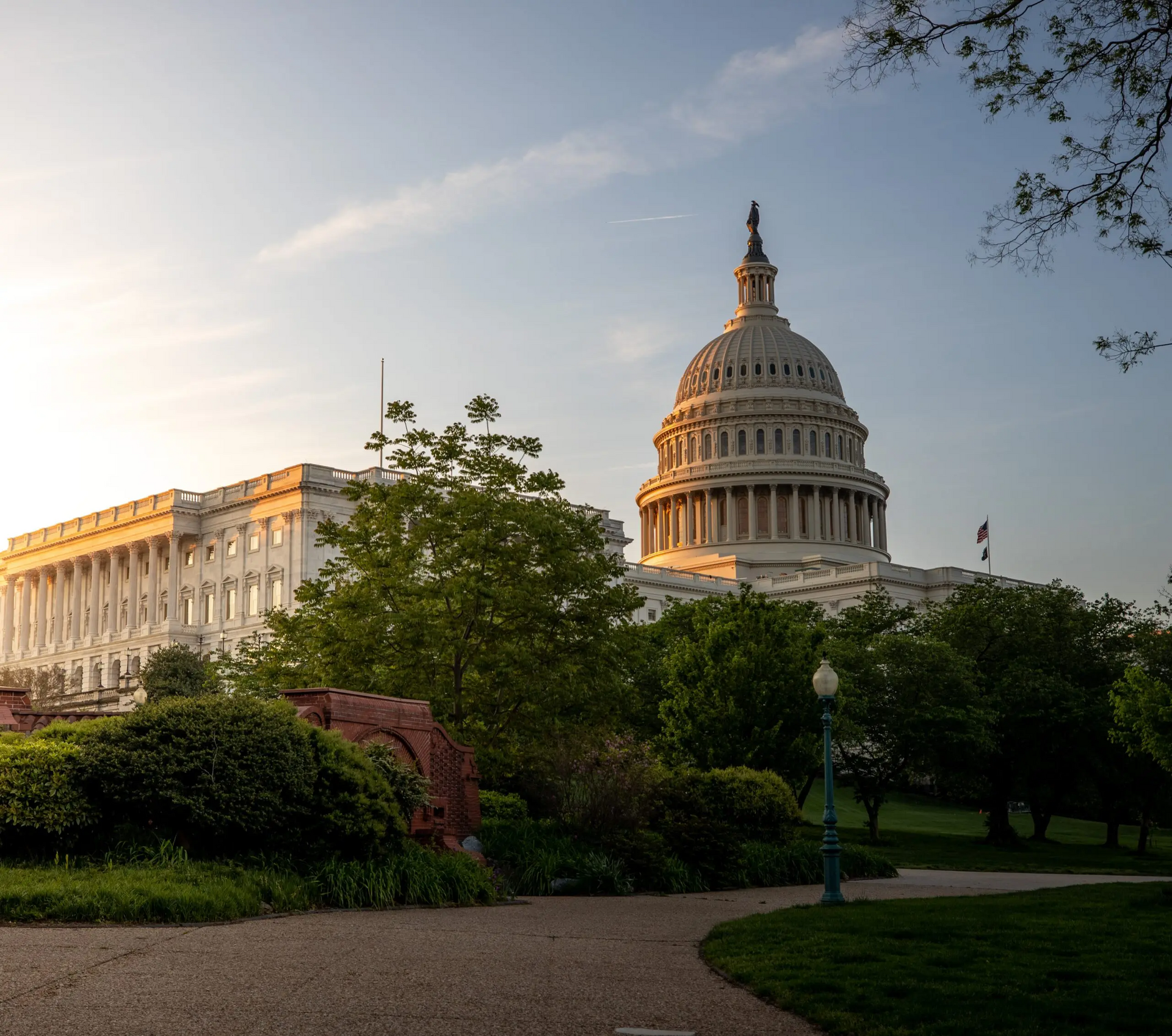 United States Capitol building bathed in sunlight, seen from a park with trees and a winding path.