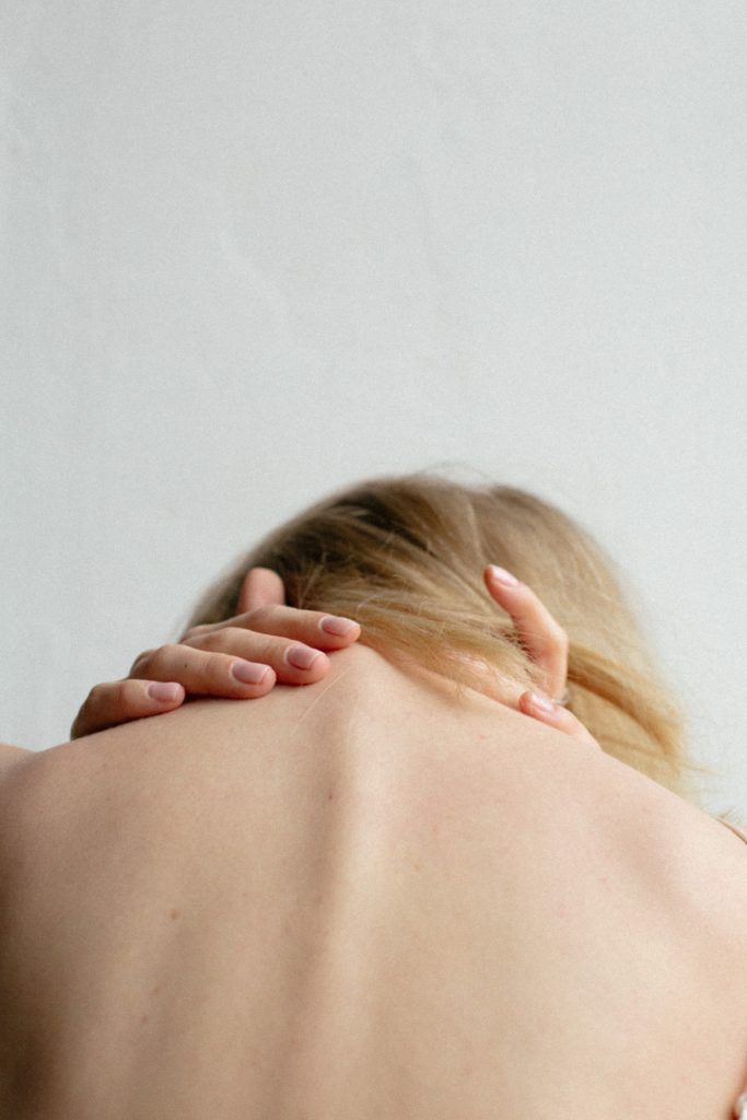 Woman with blonde hair, seen from the back, holding her neck, likely in pain, against a plain white background.