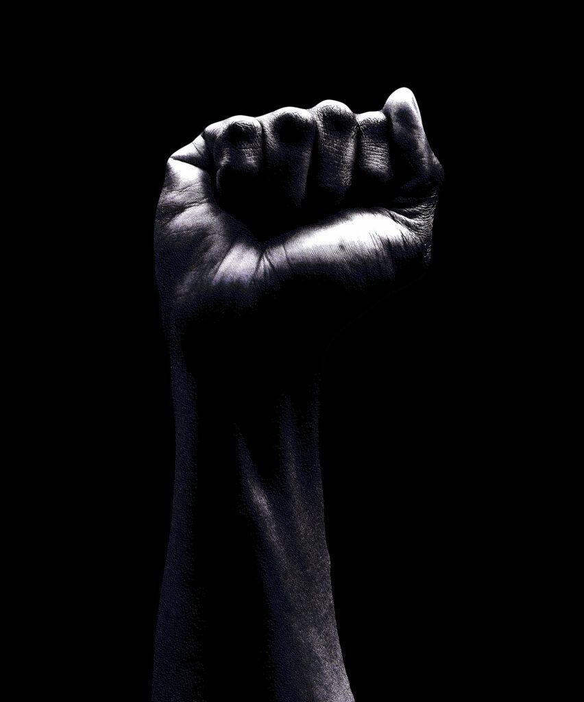 Raised fist against a black background. A hand is clenched, showing determination and solidarity.