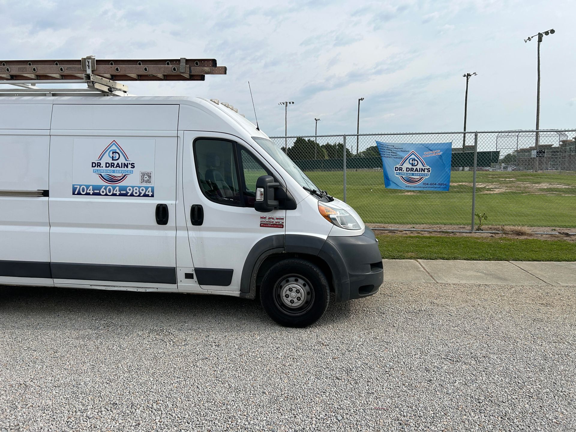 White work van with ladder parked on gravel next to a baseball field. Blue company banner in background.
