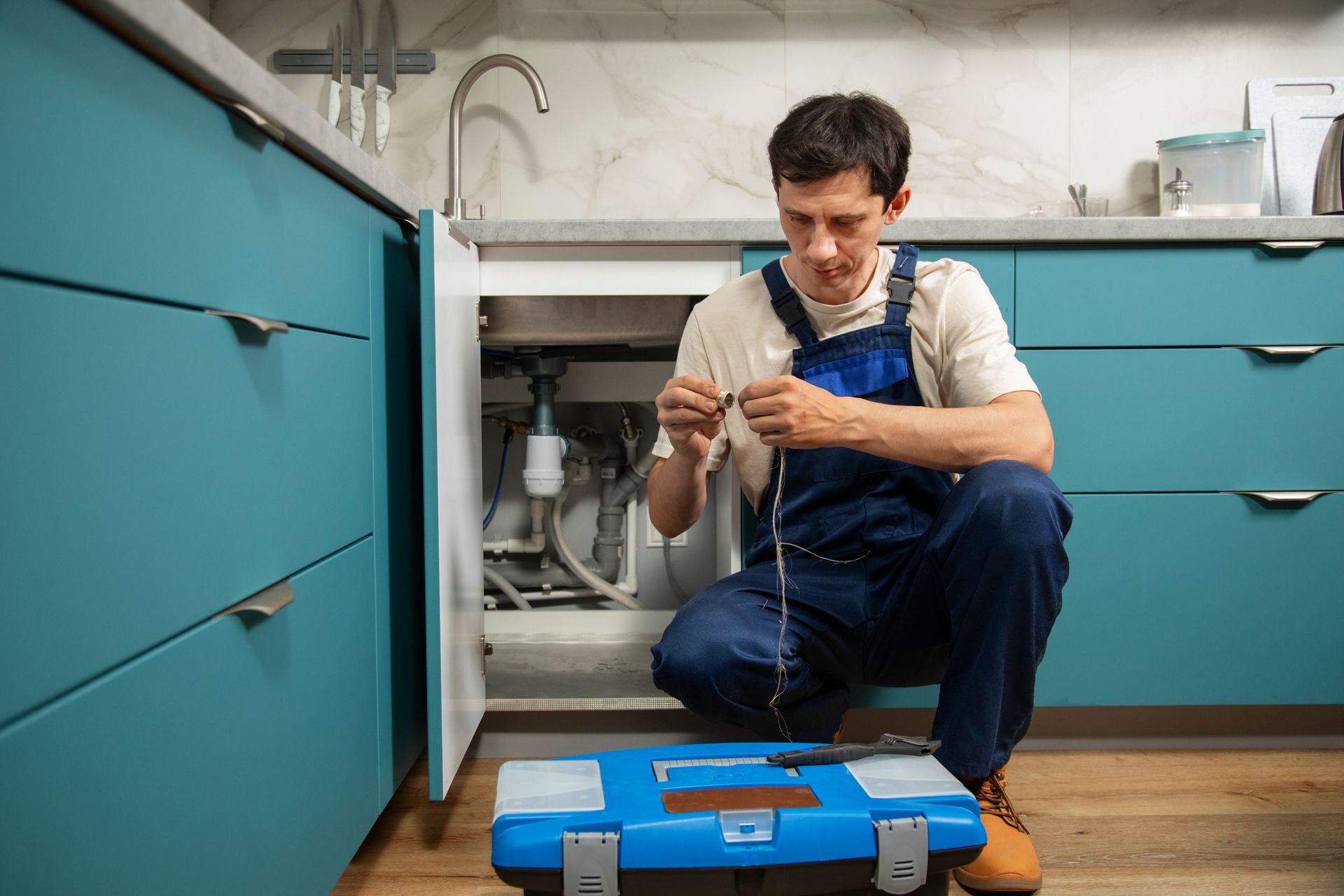Technician kneeling under a sink, repairing a pipe with a blue toolbox in a kitchen