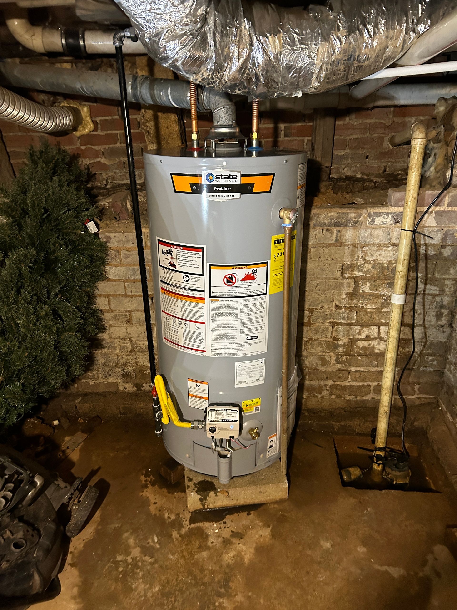 A water heater is sitting in a basement next to a brick wall.
