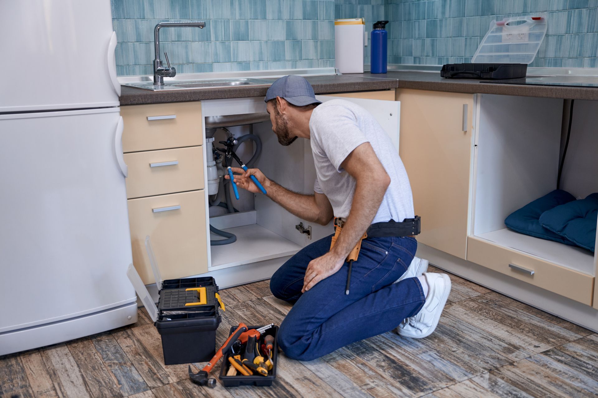 Plumber kneeling under kitchen sink, working on pipes with tools.