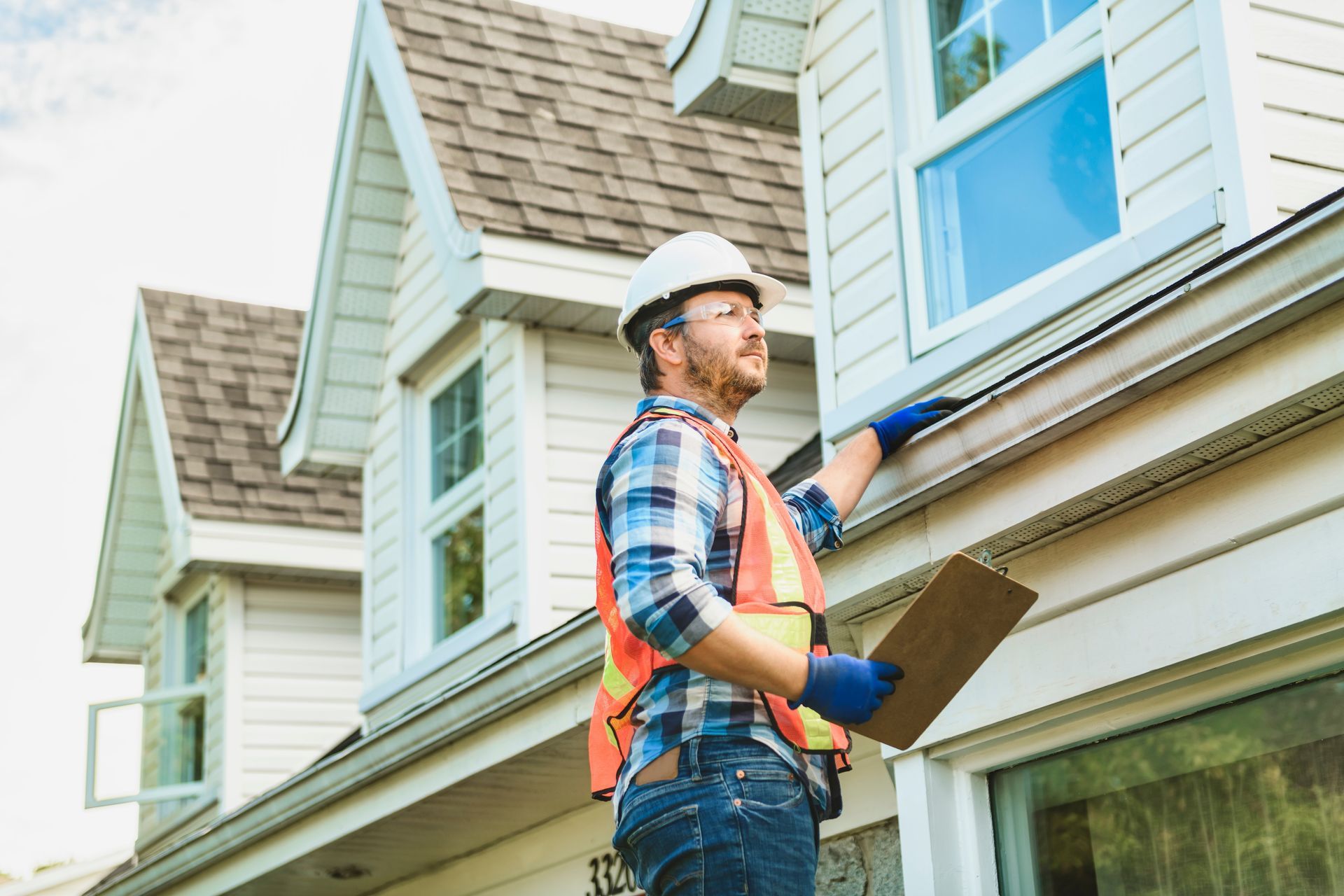 Worker in hard hat and orange vest painting a house exterior with a brush and ladder nearby