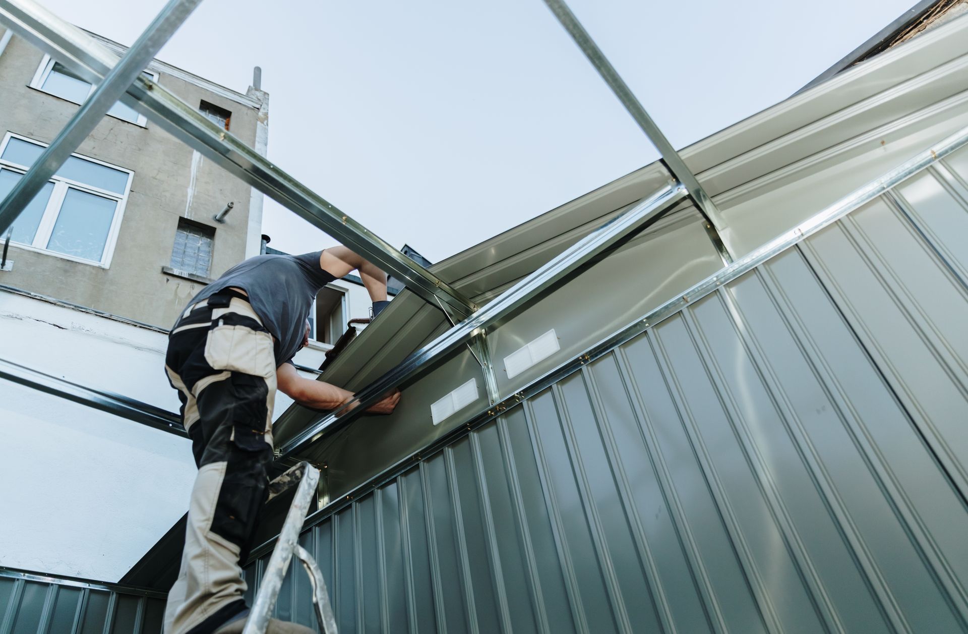 Worker on a ladder installing panels on a corrugated metal building roof.