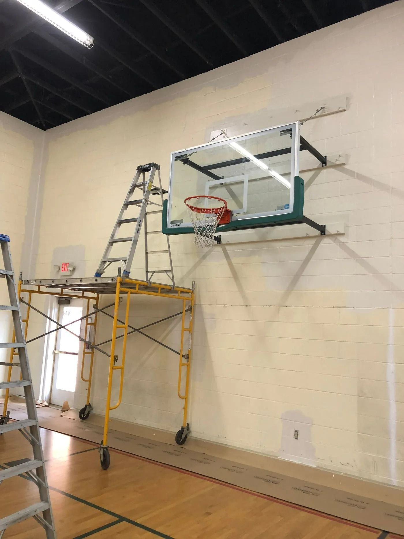 Basketball hoop mounted on a wall with a scaffolding and ladder. Wooden floor.