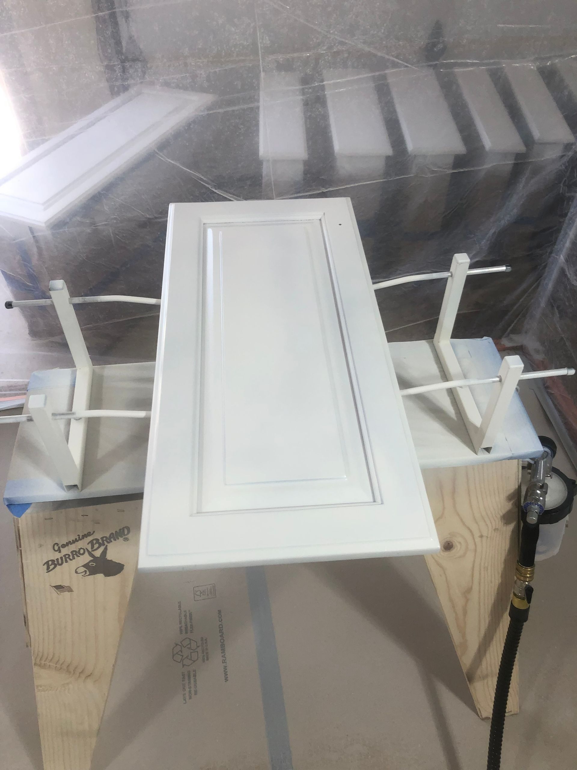 White cabinet door being spray-painted, inside a clear plastic enclosure, with other doors drying in the background.