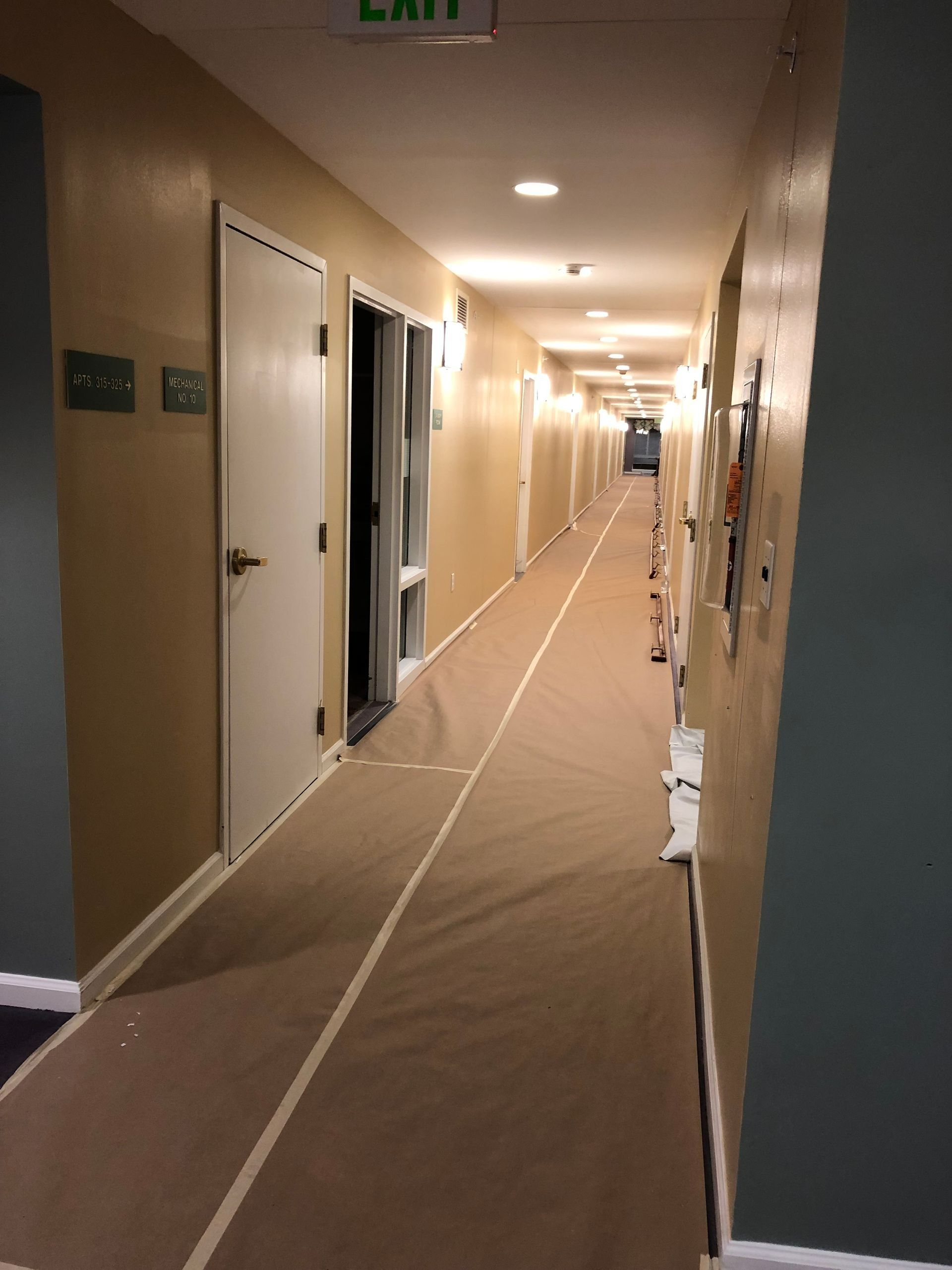 Long hallway with beige carpet, white doors, and tan walls, leading to an exit sign.